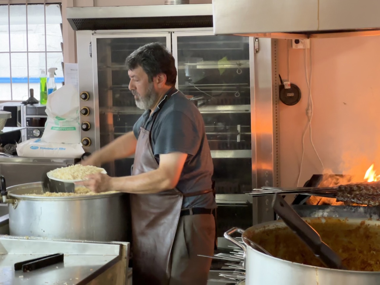 In Afghan Restaurant there are two Afghani guys with a big pot of rice and many skewers of meat grilling